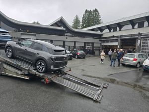 Den nye bilen vår (den grå) på veg nedover rampa på transportbilen utanfor bilverkstadhallen på skulen. Ei gruppe elevar og vaksne følger med i bakgrunnen