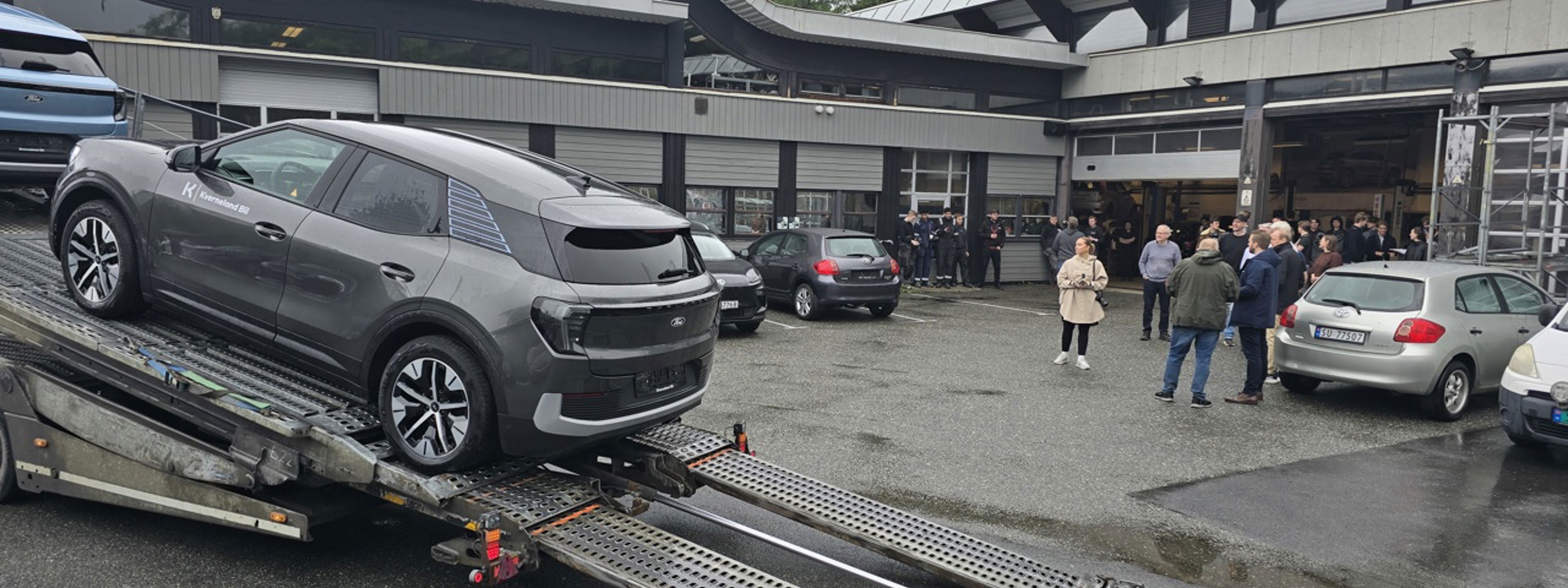 Den nye bilen vår (den grå) på veg nedover rampa på transportbilen utanfor bilverkstadhallen på skulen. Ei gruppe elevar og vaksne følger med i bakgrunnen