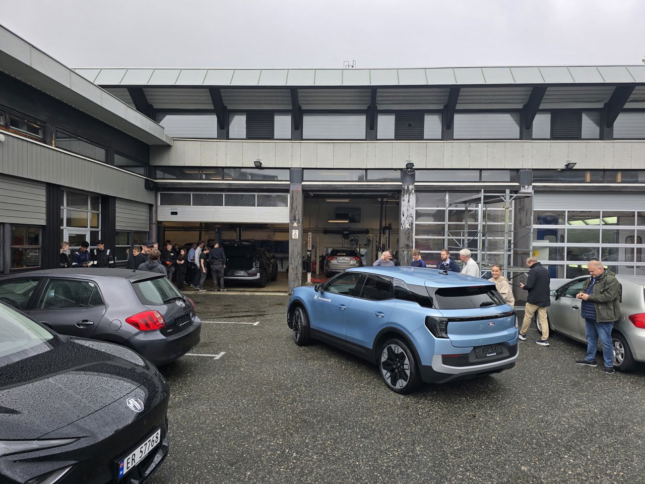 Ein av bilane (den blå) frå Kverneland BIl på plassen utanfor verkstadhallen. I bakgrunnen, inne i verkstadhallen, skimtar vi vår nye bil (den grå). Begge bilane blir studerte av elevar og vaksne.