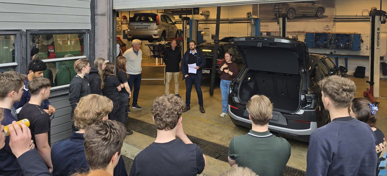 Ei gruppe elevar samla utanfor porten til bilverkstaden på skulen. Dei lyttar til ein representant frå Kverneland Bil som står rett innanfor porten, ved sida av den nye bilen.