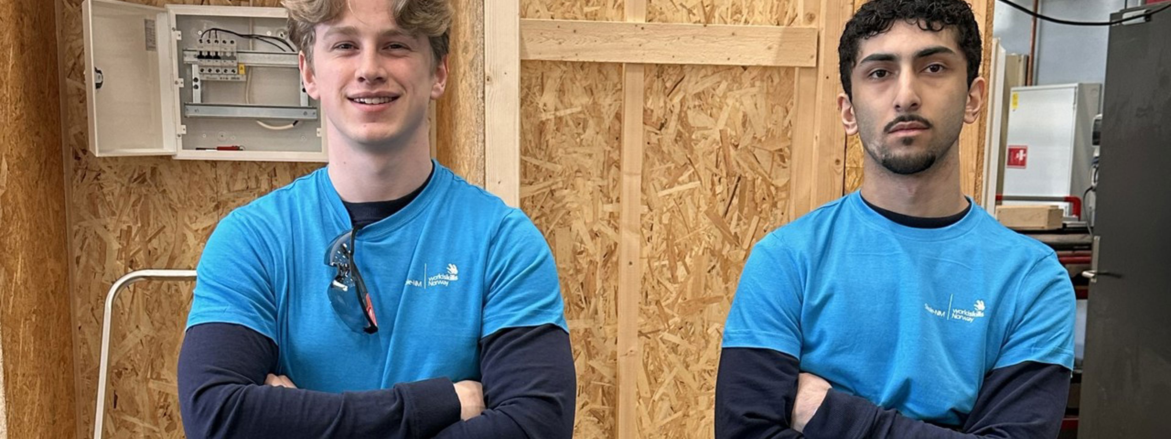 Lars og Didar, i arbeidsklede, klare for konkurranse. I bakgrunnen ein vegg med blant anna eit tomt sikringsskap.