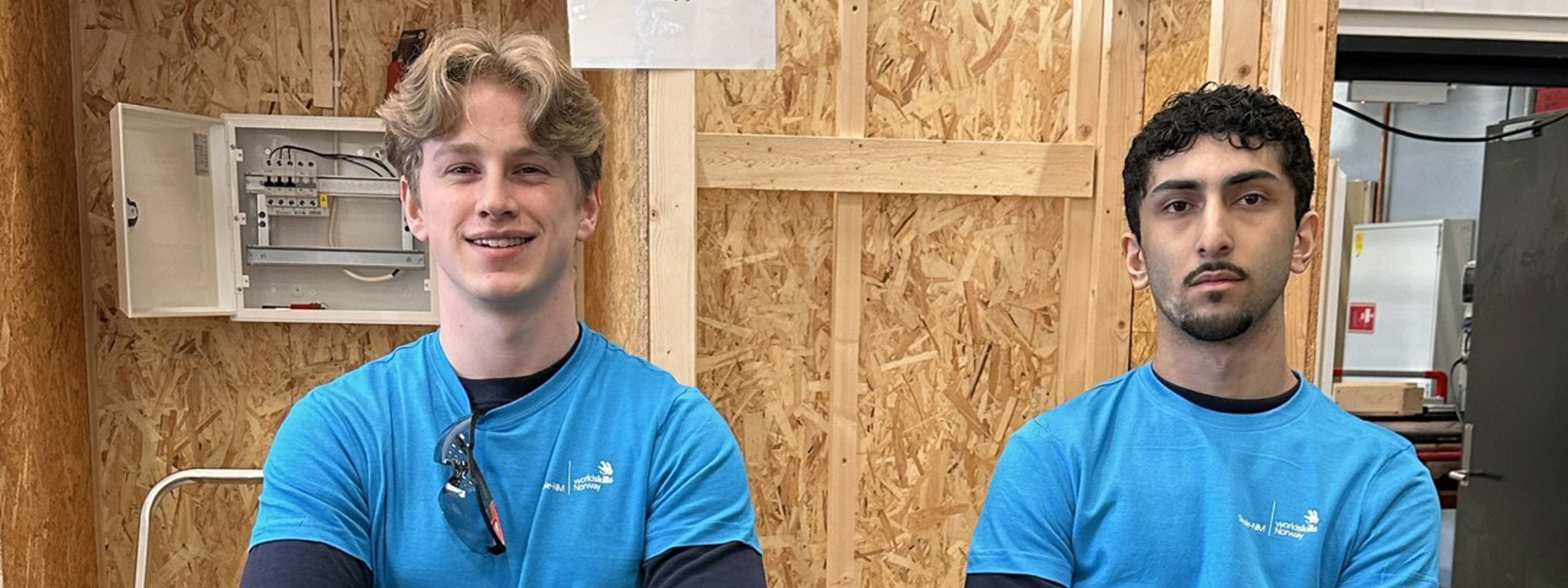 Lars og Didar, i arbeidsklede, klare for konkurranse. I bakgrunnen ein vegg med blant anna eit tomt sikringsskap.