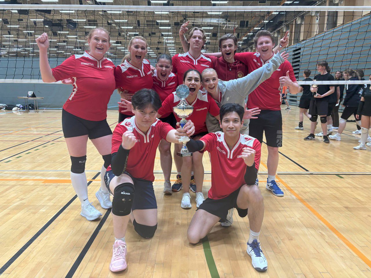 Eit jublande Mix-lag poserer med pokalen dei har vunne, foran volleyballnettet.