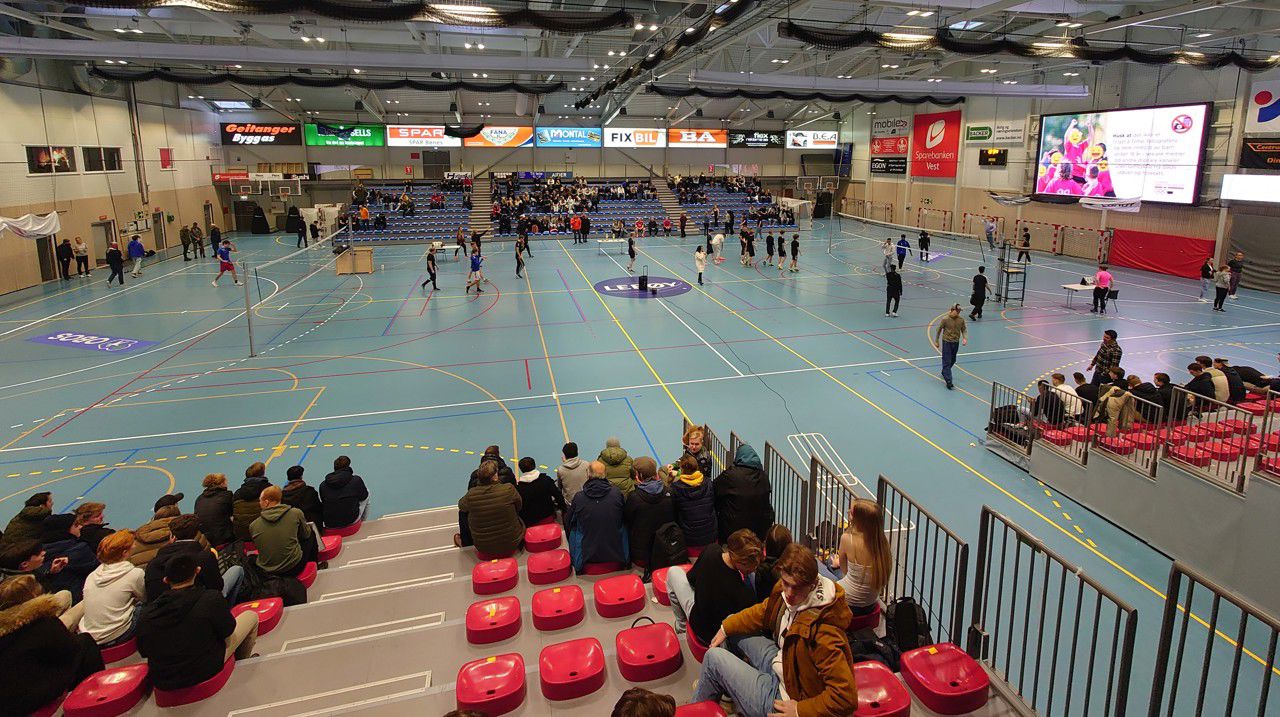 Bilde over idrettshallen, tatt frå toppen av tribunen på eine langveggen. Det er rigga til med to volleyballbanar og tribunar på begge langsidene. Ein god del folk på tribunane (men langt frå fullt). Volleyball-laga gjer seg klare på banen og, midt i det heile, ventar rektor på å ønske velkommen.