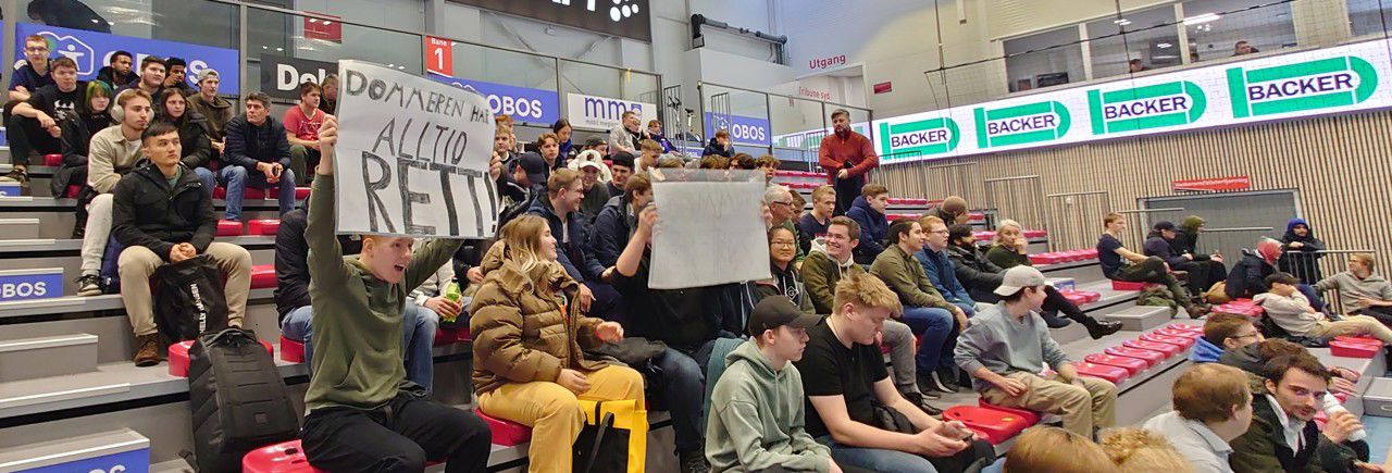 Bildet viser ei gruppe elevar og lærarar som sit på tribunen. Ein av elevane held opp ein stor plakat der det står "Dommeren har alltid rett!" med store bokstavar.