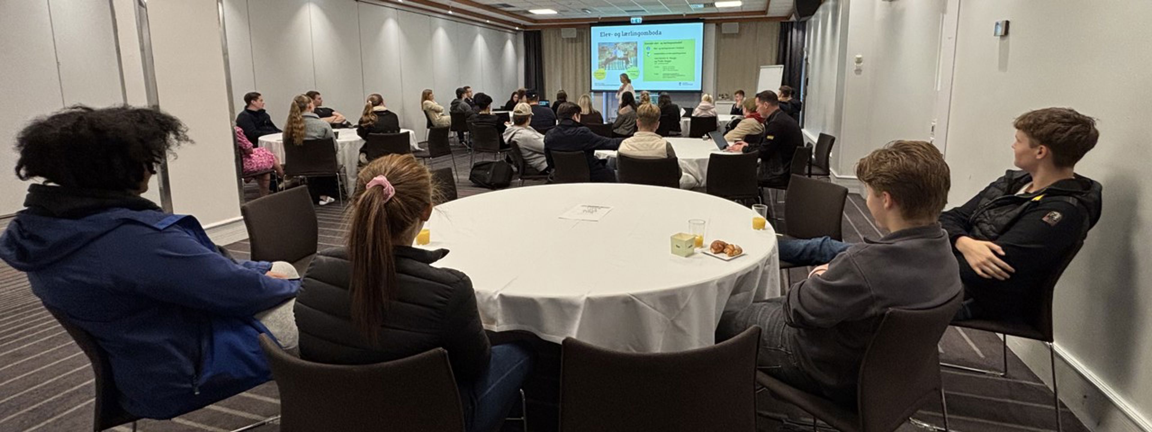Ei gruppe elevar (tillitsvalde) samla ved fleire runde konferansebord følger med på presentasjonen til elev- og lærlingomboda