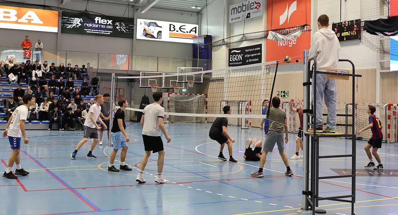 Bilde frå spelet i gutefinalen. Vi ser begge laga i farten, med dommaren på eit podium ved nettet og publikum på tribunen i bakgrunnen.