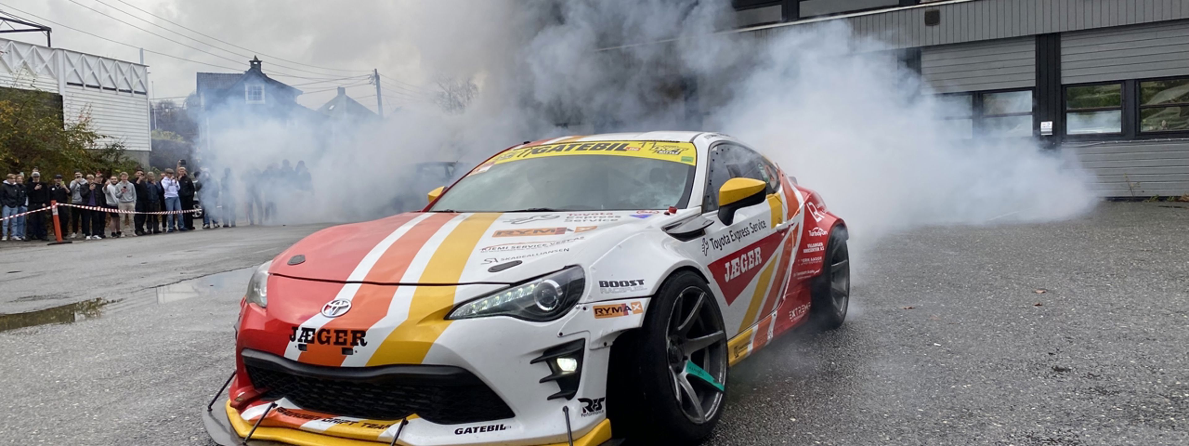 Blå røyk frå bakhjula på Davanger Drifting-bilen tåkelegg skuleplassen utanfor verkstadhallane. Publikum følger med på trygg avstand.