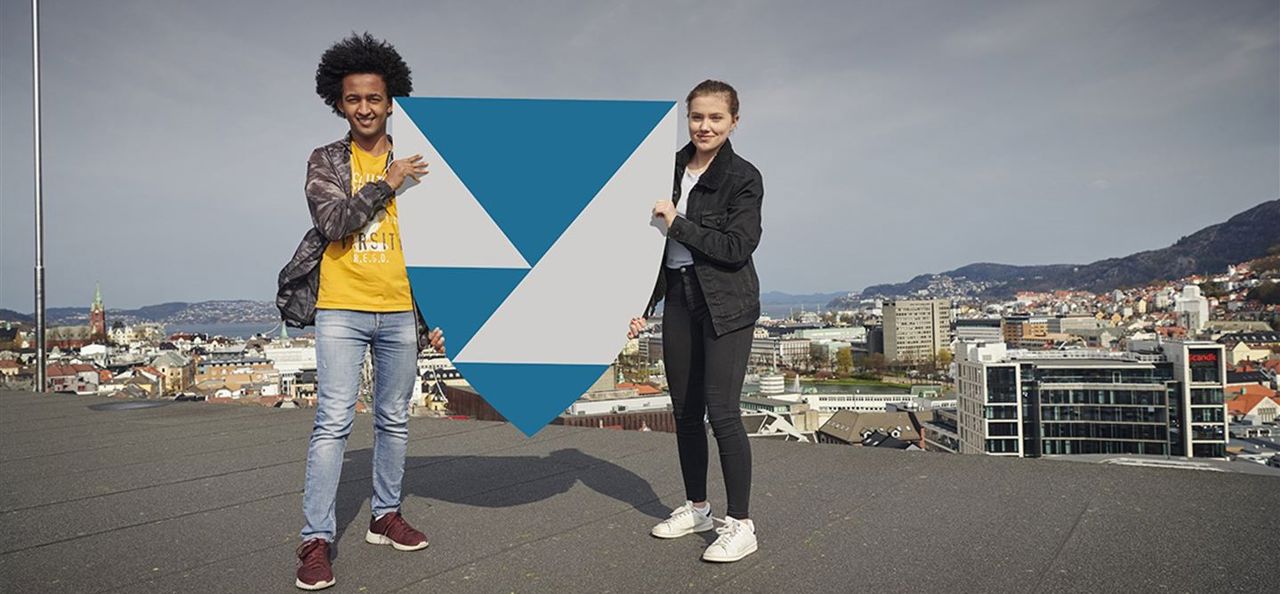 To ungdommar på taket av det gamle fylkesbygget i Bergen held ei diger utgåve av fylkesvåpenet til Vestland fylkeskommune mellom seg. 